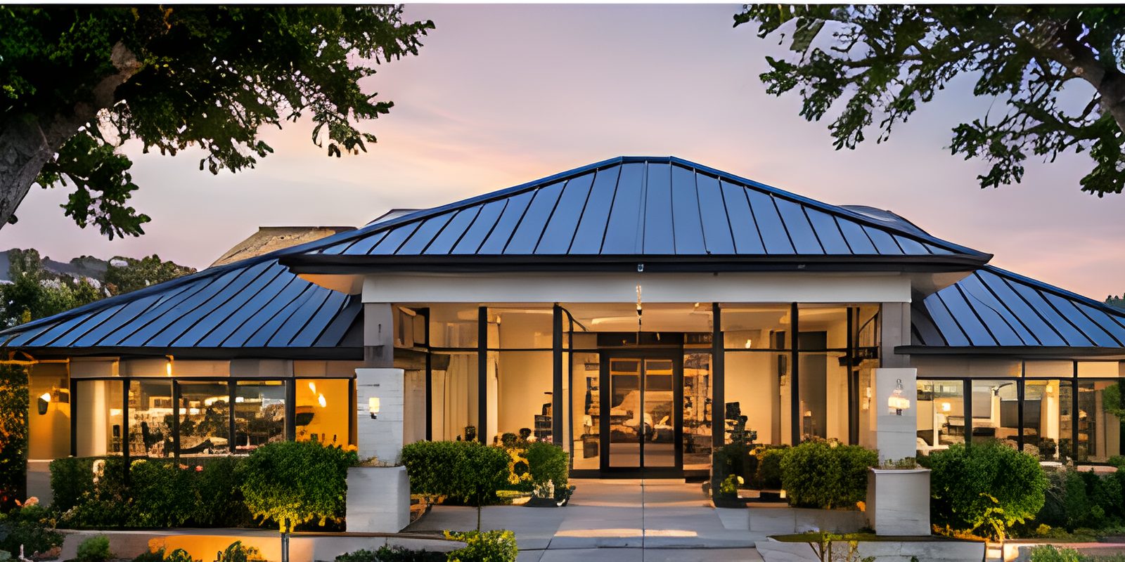 A single-storey pavilion with a standing-seam metal roof at dusk, lit from within.